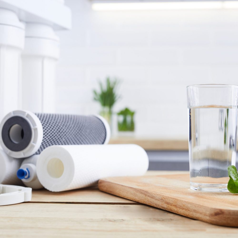 A glass of clean water with osmosis filter, green leaves and cartridges on wooden table in kitchen interior. Concept Household filtration system.